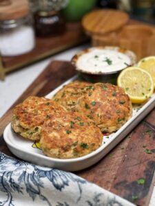 A white rectangular platter holding four canned tuna patties that have been sautéed and browned in olive oil. The patties are served with a bowl of lemon dill sauce and lemon wedges.