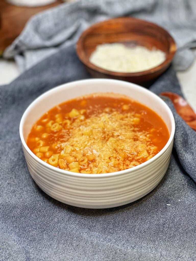 A white bowl of Italian Pasta Beans. The tomato based dish is filled with white beans and a ditalini pasta. The dish is topped with freshly grated Parmesan cheese.