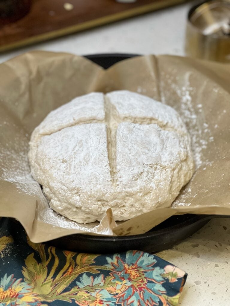A round of dough with a cross pattern cut out on top. The dough is sitting on a piece of parchment paper in a cast iron pan. 