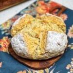 A round loaf of Easy Irish Soda Bread. The loaf was dusted with rice flour before baking and has a golden crust. It's sitting on a small wooden board.