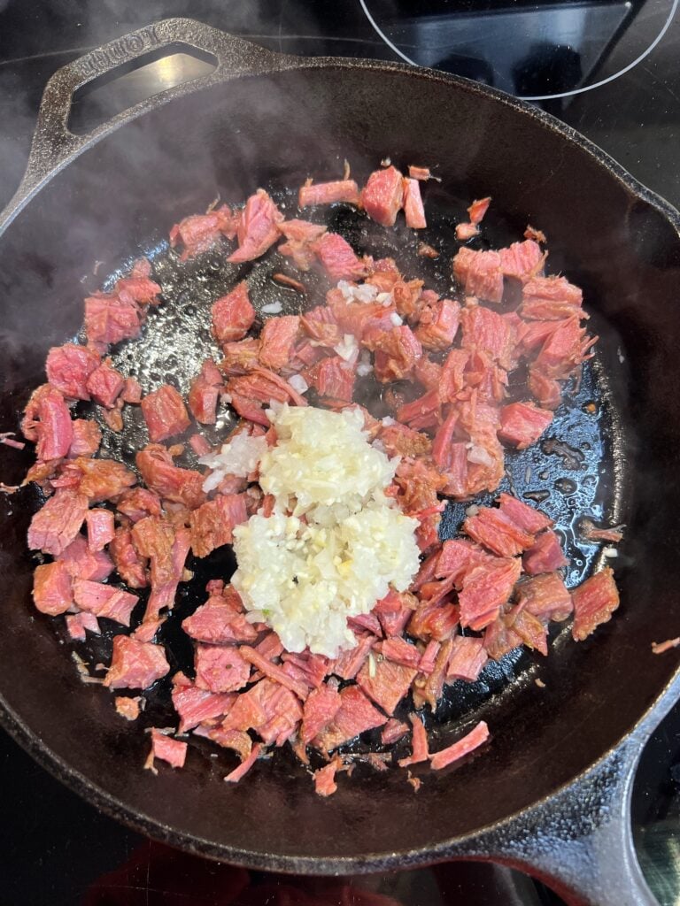 Diced corned beef crisping in a cast iron pan with minced garlic and onions.