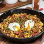 A cast iron pan of Crispy Corned Beef Hash. The has is topped with two poached eggs and garnished with fresh parsley. The hash contains crispy diced corned beef and crispy diced potatoes.