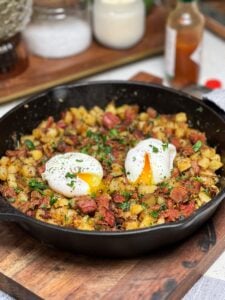 A cast iron pan of Crispy Corned Beef Hash. The has is topped with two poached eggs and garnished with fresh parsley. The hash contains crispy diced corned beef and crispy diced potatoes.