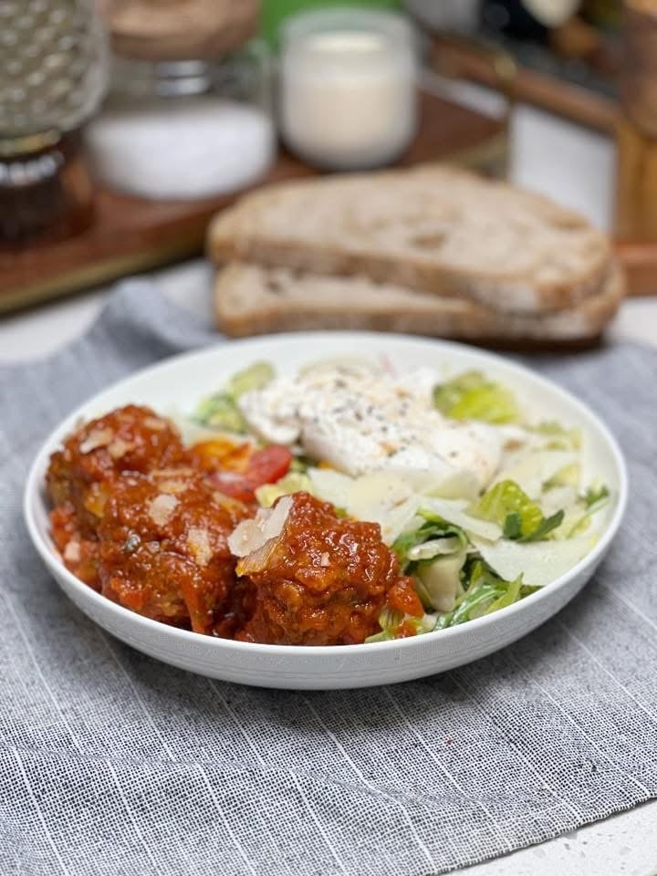 A meatball salad with simple vinaigrette. A bowl of salad topped with an open piece of burrata with three meatballs.