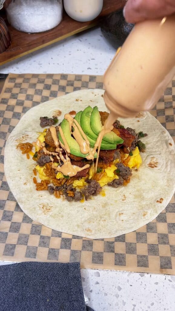 A floured tortilla filled in the center with cheese, eggs, bacon, sausage, avocado slices, and rice. The picture is showing drizzling chipotle mayo out of a squeeze bottle onto the ingredients. This is for a loaded breakfast burrito.