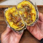 A loaded breakfast burrito wrapped in parchment and foil. The photo is showing what the burrito looks like when cut in half. You can see the flour tortilla, scrambled eggs, sausage, bacon, cheese, rice and avocado.