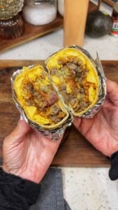 A loaded breakfast burrito wrapped in parchment and foil. The photo is showing what the burrito looks like when cut in half. You can see the flour tortilla, scrambled eggs, sausage, bacon, cheese, rice and avocado.