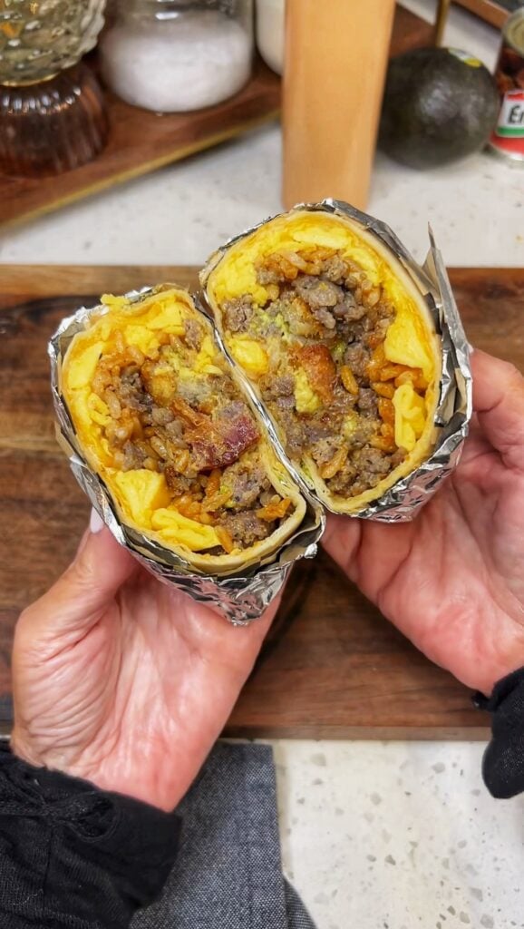 A loaded breakfast burrito wrapped in parchment and foil. The photo is showing what the burrito looks like when cut in half. You can see the flour tortilla, scrambled eggs, sausage, bacon, cheese, rice and avocado.