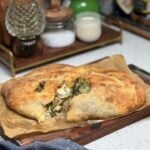 A mushroom cheese calzone cut in half sitting on a piece of brown parchment paper on a wooden board. The calzone is browned and you can see the melty cheese, mushrooms and baby argula.