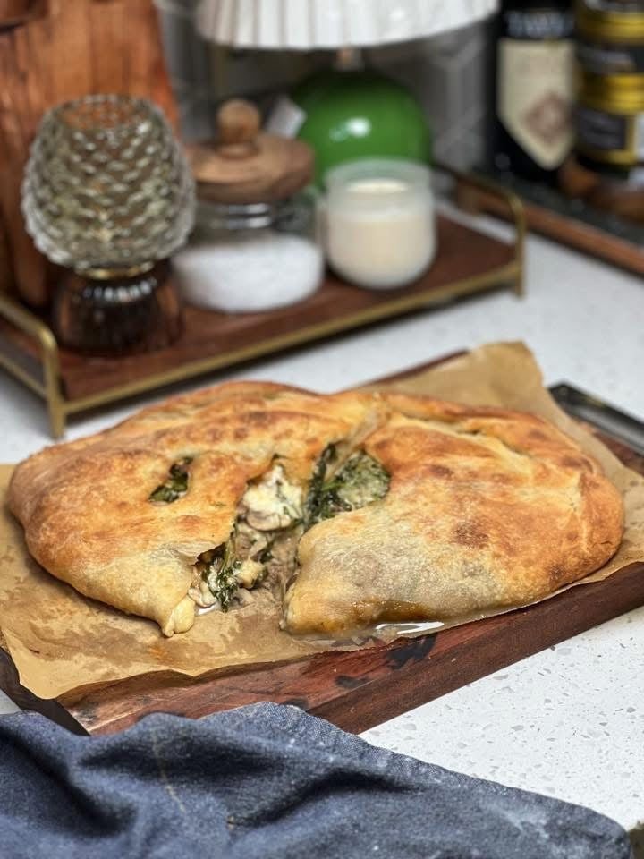 A mushroom cheese calzone cut in half sitting on a piece of brown parchment paper on a wooden board. The calzone is browned and you can see the melty cheese, mushrooms and baby argula.