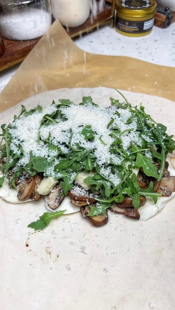 One half of a rolled out pizza dough holding fresh mozzarella slices, sautéed mushrooms, pieces of fontina cheese, arugula and grated Parmesan cheese.