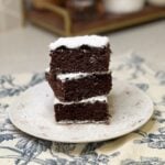 Three pieces of sheet pan chocolate depression cake cut into squares and dusted with powdered sugar. The three pieces are stacked on top of each other on a white plate.