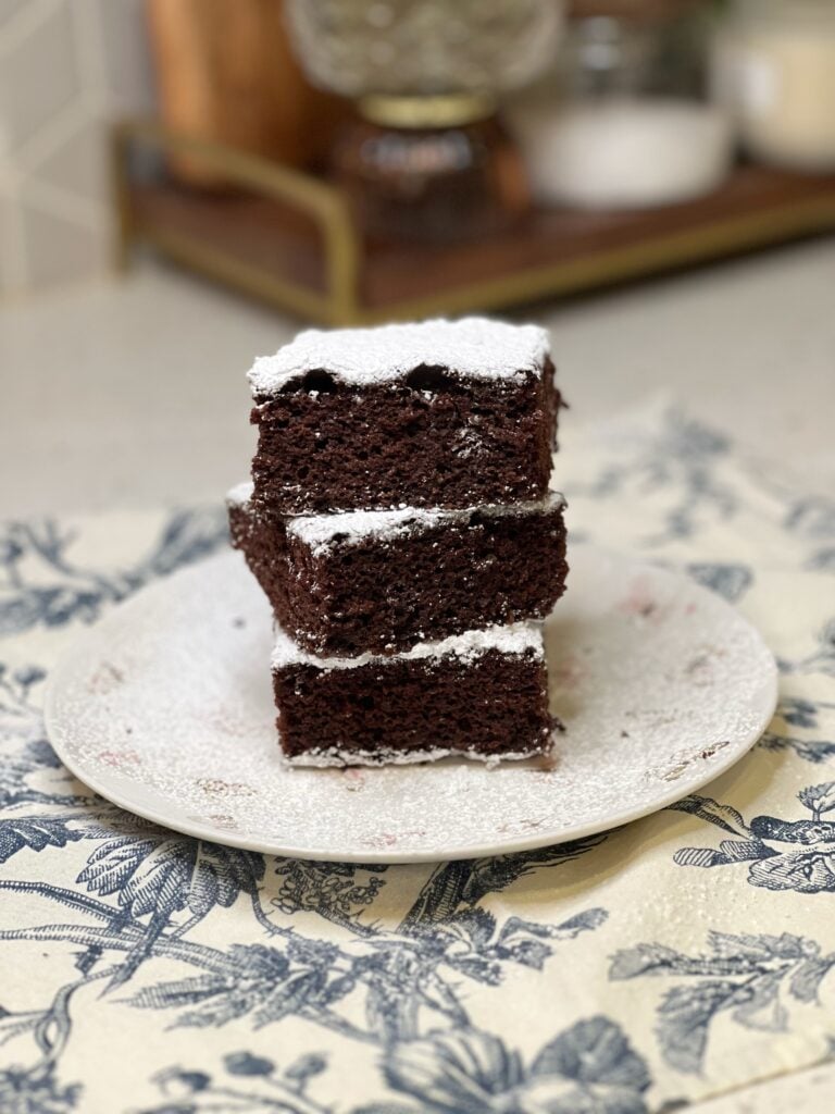 Three pieces of sheet pan chocolate depression cake cut into squares and dusted with powdered sugar. The three pieces are stacked on top of each other on a white plate.
