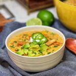 A bowl of creamy taco soup. The soup has an orangish hue with tomatoes, black beans, shredded chicken and rice. The soup is garnished with tortilla strips, diced avocado, sour cream and sliced jalapeños.
