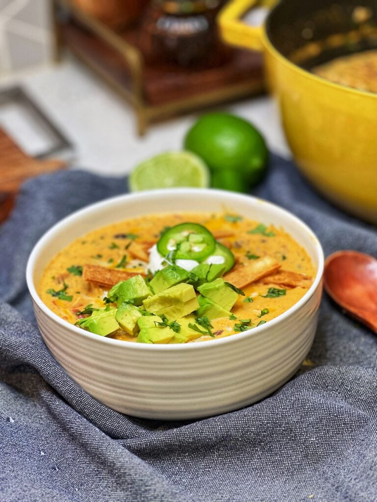 A bowl of creamy taco soup. The soup has an orangish hue with tomatoes, black beans, shredded chicken and rice. The soup is garnished with tortilla strips, diced avocado, sour cream and sliced jalapeños.