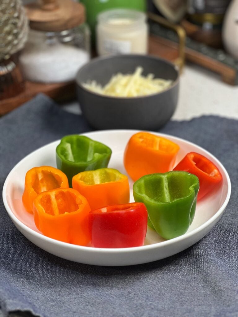 Eight mini bell peppers in a variety of colors sitting in a white bowl. The peppers have their tops cut off and the seeds and ribs removed.