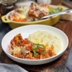Two mini stuffed bell peppers in a white bowl with a side of mashed potatoes. The peppers are covered in a meat sauce and melted mozzarella cheese.