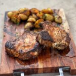Two juicy grilled pork chops on a wooden board with grilled potatoes in the background. The chops are about an inch thick with the bone-in.