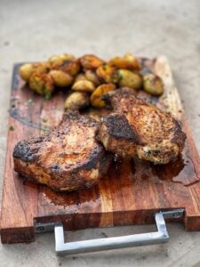 Two juicy grilled pork chops on a wooden board with grilled potatoes in the background. The chops are about an inch thick with the bone-in.