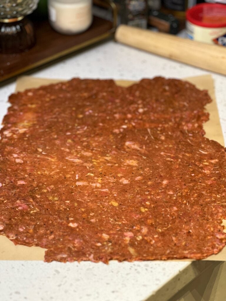 Ground meat and spices mixed together and flattened thin on a piece of brown parchment paper.