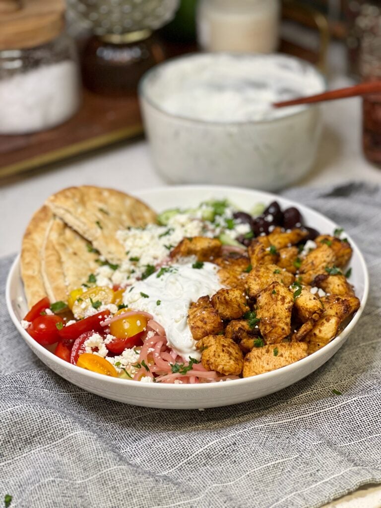An air fryer Greek chicken bowl with pita, onions, tomatoes, feta cheese, cucumbers, tzatziki sauce and olives.