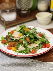 A plate of Pork Milanese with Arugula Salad. Crispy fried pork is topped with an arugula salad with cherry tomatoes and shaved Parmesan cheese.