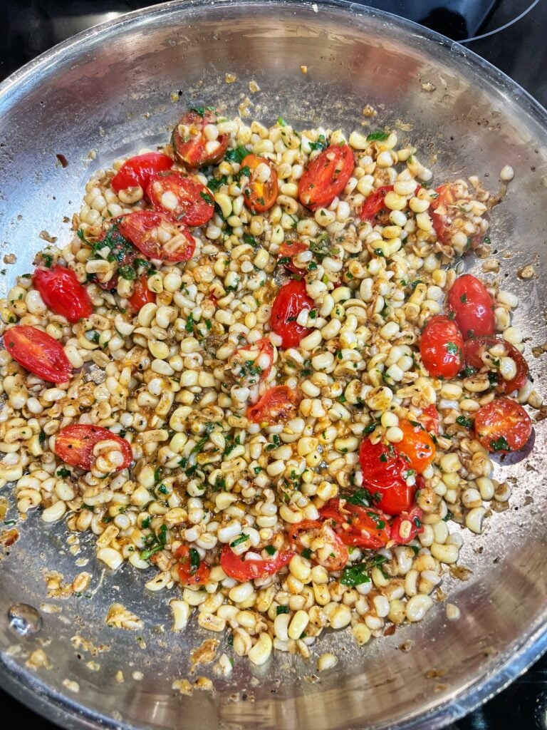 A stainless steel pan of brown butter sweet corn with blistered tomatoes and fresh parsley.