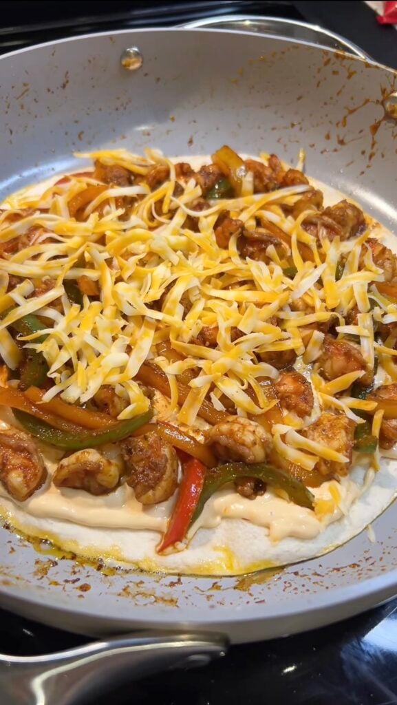 A large flour tortilla in a non-stick pan filled with a creamy cheesy layer, sautéed shrimp and peppers and topped with shredded cheese for shrimp fajita quesadillas.