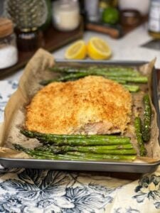 A filet of baked panko crusted salmon with asparagus on a parchment-lined baking sheet. The salmon has a browned crust.