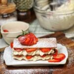 A strawberry shortcake napoleon. Rectangular strips of baked pastry crust are layered with mascarpone cream and fresh sliced strawberries. The napoleon is sprinkled with powdered sugar and garnished with a fresh, fanned out strawberry.