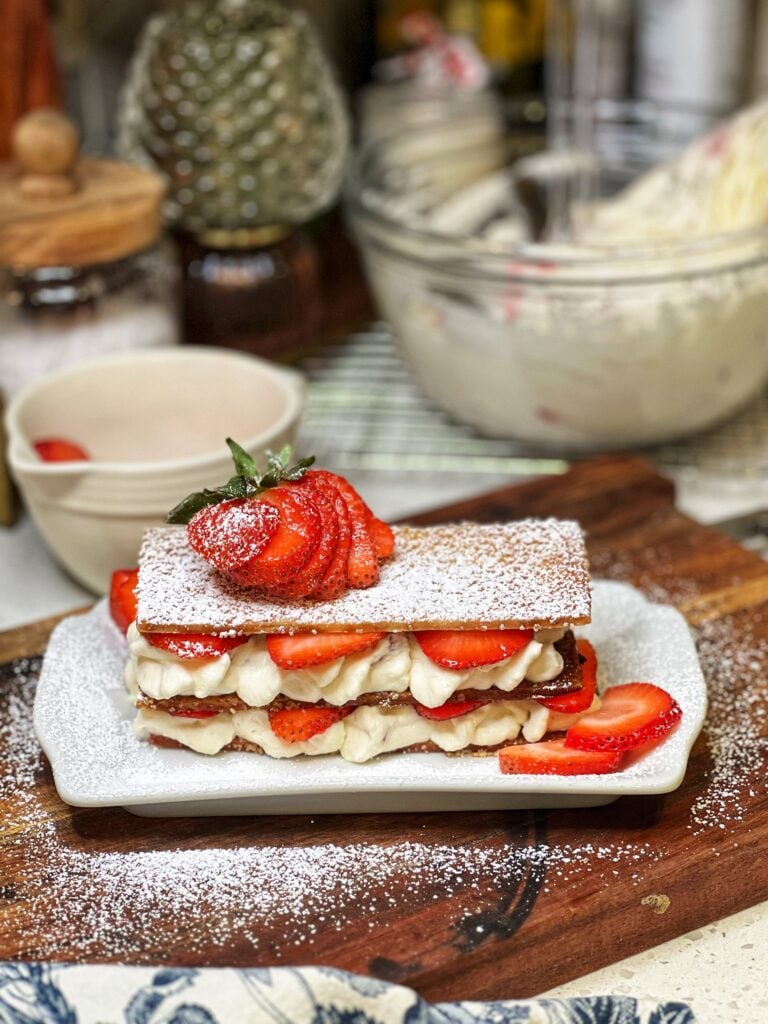 A strawberry shortcake napoleon. Rectangular strips of baked pastry crust are layered with mascarpone cream and fresh sliced strawberries. The napoleon is sprinkled with powdered sugar and garnished with a fresh, fanned out strawberry.