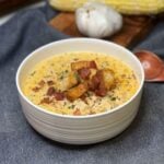 A white bowl of chicken corn chowder that is garnished with homemade croutons, crisp bacon pieces, fresh thyme and cracked black pepper.