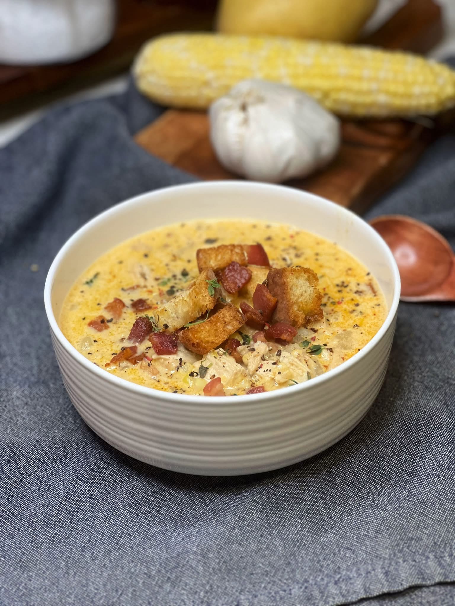 A white bowl of chicken corn chowder that is garnished with homemade croutons, crisp bacon pieces, fresh thyme and cracked black pepper.