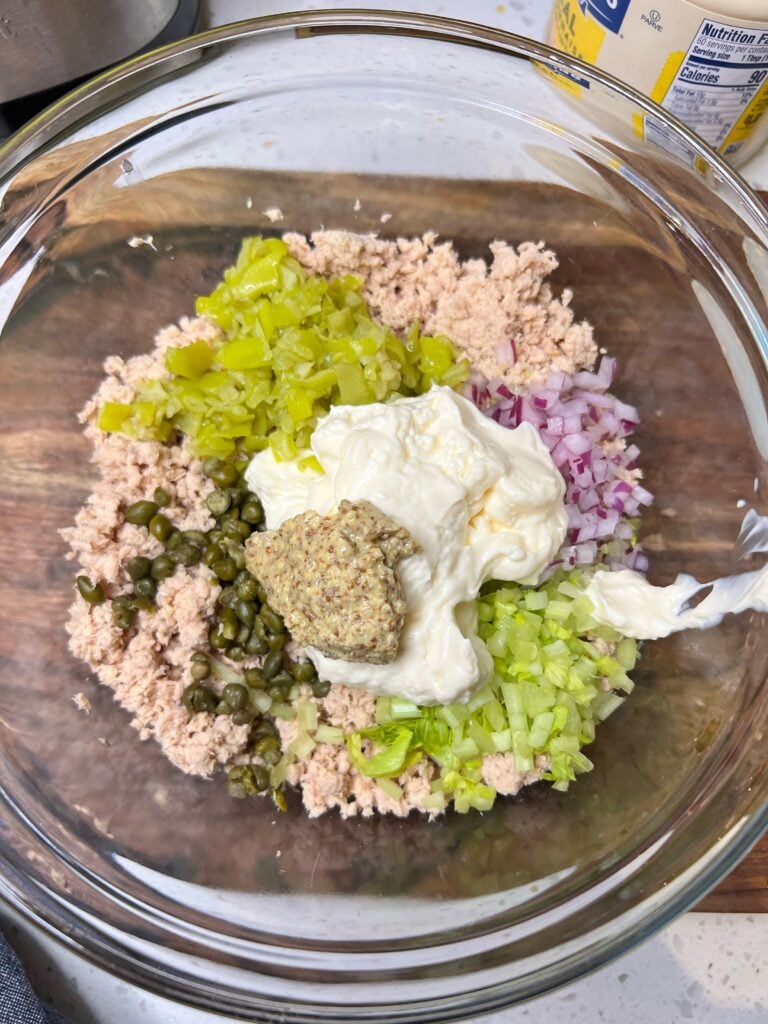 Ingredients in a glass mixing bowl for pepperoncini tuna salad. The bowl contains canned tuna, celery, red onion, peppers, capers, mayo and Dijon mustard.