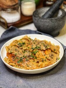 A white bowl filled with Peppercorn Chicken Pasta with Mushrooms. Pappardelle pasta is in a creamy mushrooms sauce topped with two seared chicken breasts and garnished with fresh parsley.