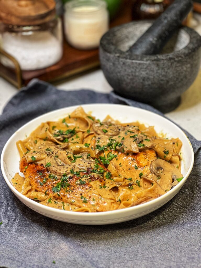 A white bowl filled with Peppercorn Chicken Pasta with Mushrooms. Pappardelle pasta is in a creamy mushrooms sauce topped with two seared chicken breasts and garnished with fresh parsley.