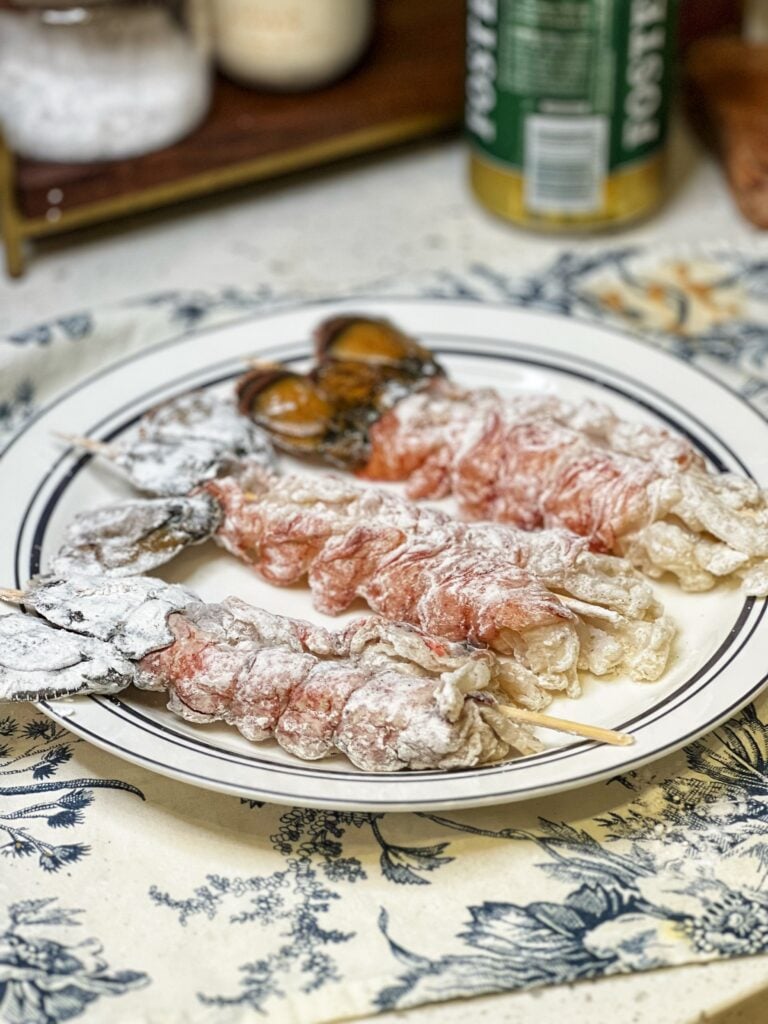 Three raw lobster tails coated in seasoned flour sitting on a plate.