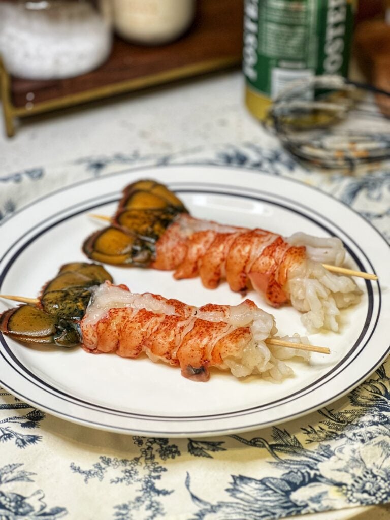 Two raw lobster tails on a plate, each with a wooden skewer lengthwise in the tail.
