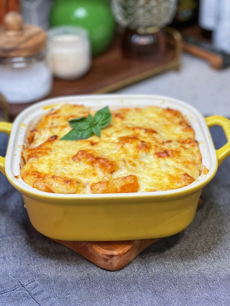 A yellow, square casserole dish filled with Baked Rigatoni with Meat Sauce. The top of the casserole has a browned cheesy crust, and is garnished with fresh basil.
