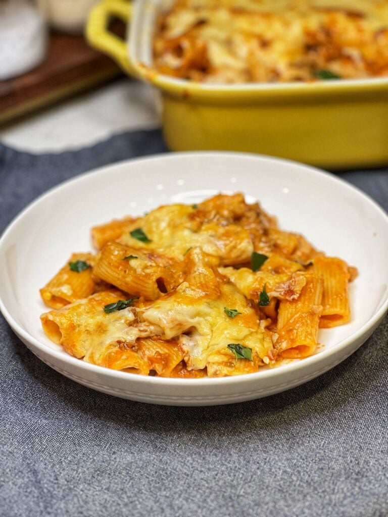 A white, shallow bowl filled with a few spoonfuls of baked rigatoni in a meat and béchamel sauce that has browned cheese on top. The dish is garnished with fresh basil.