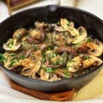 A cast iron pan of oven roasted butter garlic mushrooms. The large mushrooms are halved in a butter garlic sauce and garnished with fresh chopped parsley.
