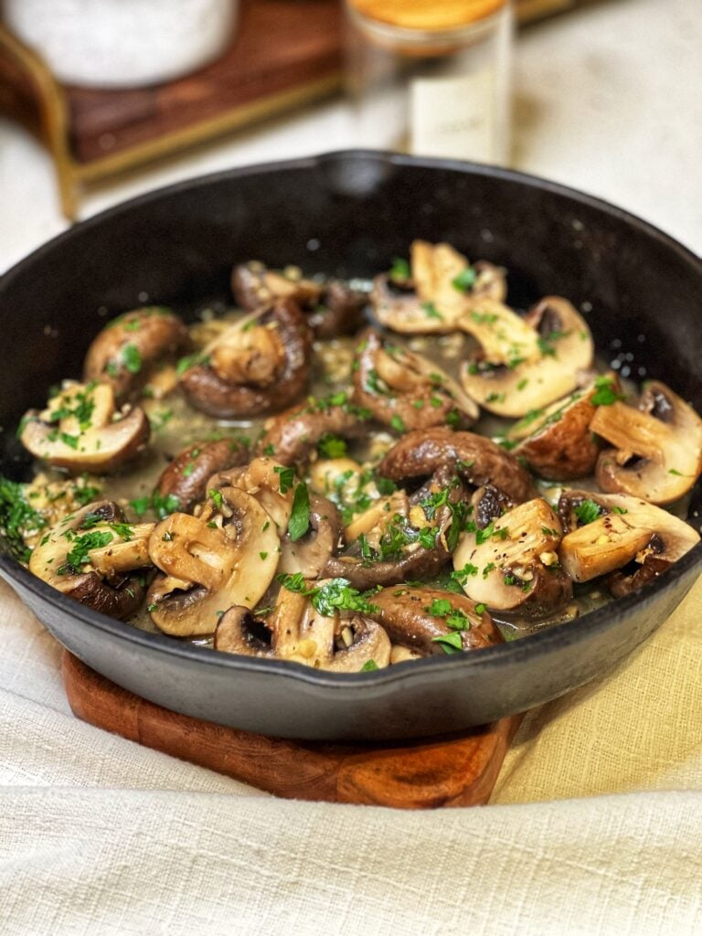 A cast iron pan of oven roasted butter garlic mushrooms. The large mushrooms are halved in a butter garlic sauce and garnished with fresh chopped parsley.