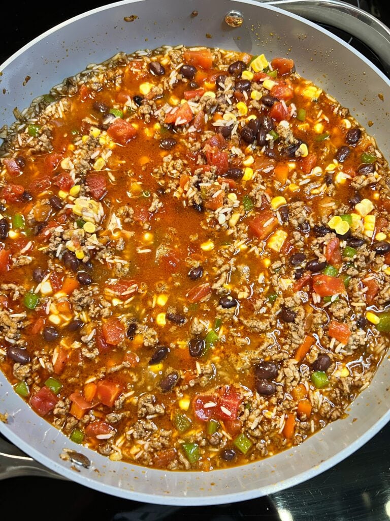 A skillet of browned ground beef simmering with rice, diced peppers, tomatoes, corn, black beans in beef broth.