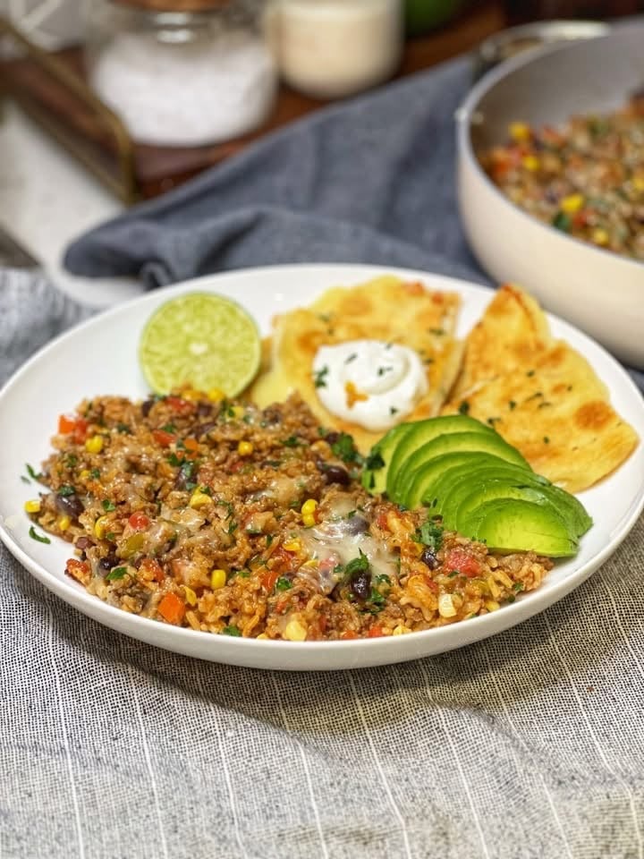 A shallow bowl with a few spoonfuls of beef taco rice, sliced avocado, a lime wedge, and triangles of cheese quesadillas topped with sour cream.