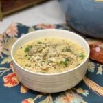 A white bowl filled with homemade chicken and dumplings. The chicken is shredded in a rich, creamy broth with flat dumplings. The dish is garnished with fresh parsley and black peppr.