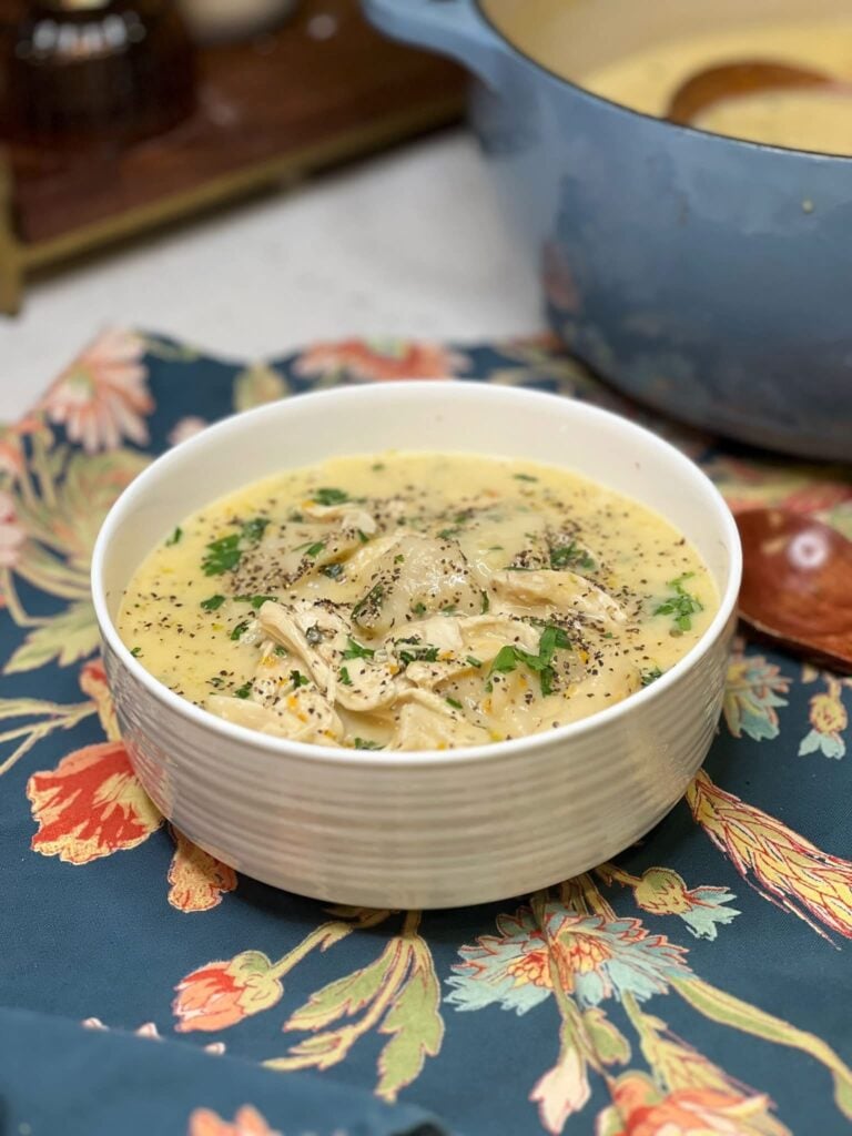 A white bowl filled with homemade chicken and dumplings. The chicken is shredded in a rich, creamy broth with flat dumplings. The dish is garnished with fresh parsley and black peppr.