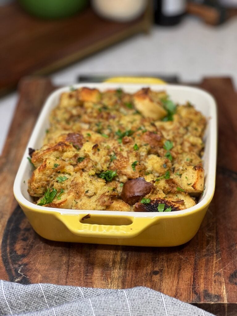 A casserole dish of baked, homemade herb stuffing. The top is browned and garnished with fresh parsley.