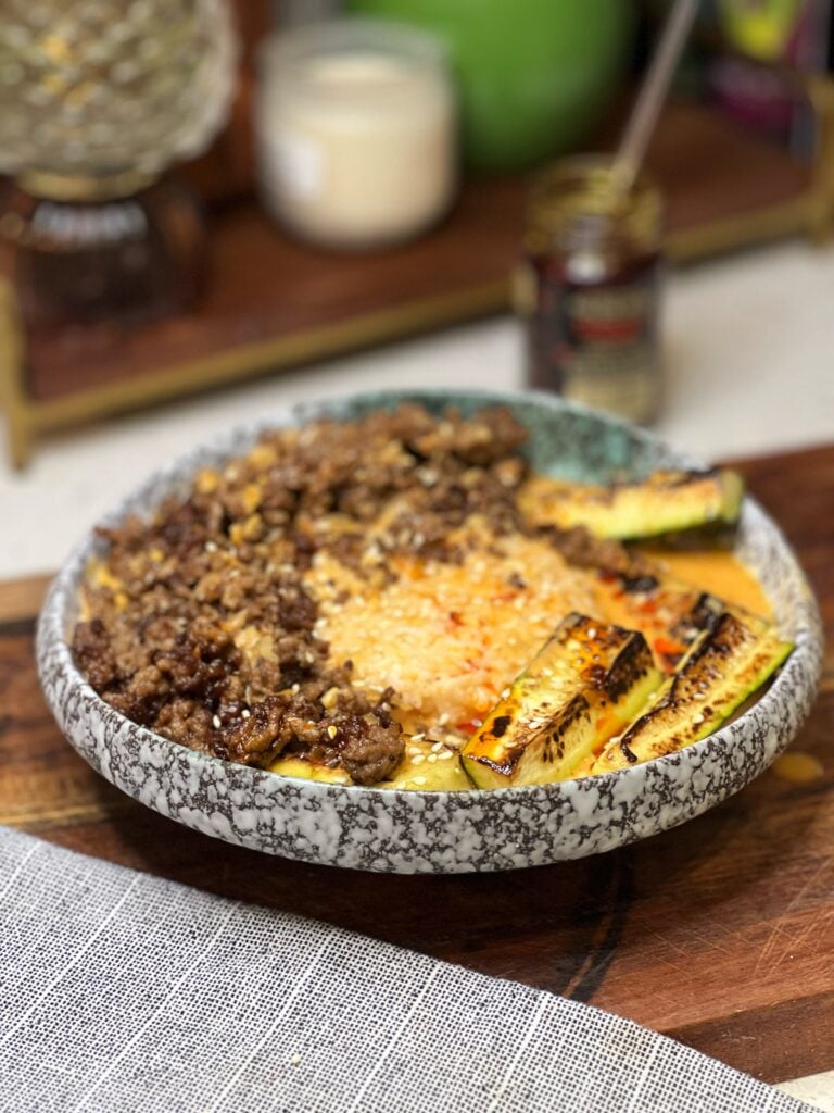 A Curry Beef Broth Bowl. A mound of steamed rice is served with crispy ground beef and seared zucchini spears on the side with a coconut curry broth on top and a drizzle of chili oil.